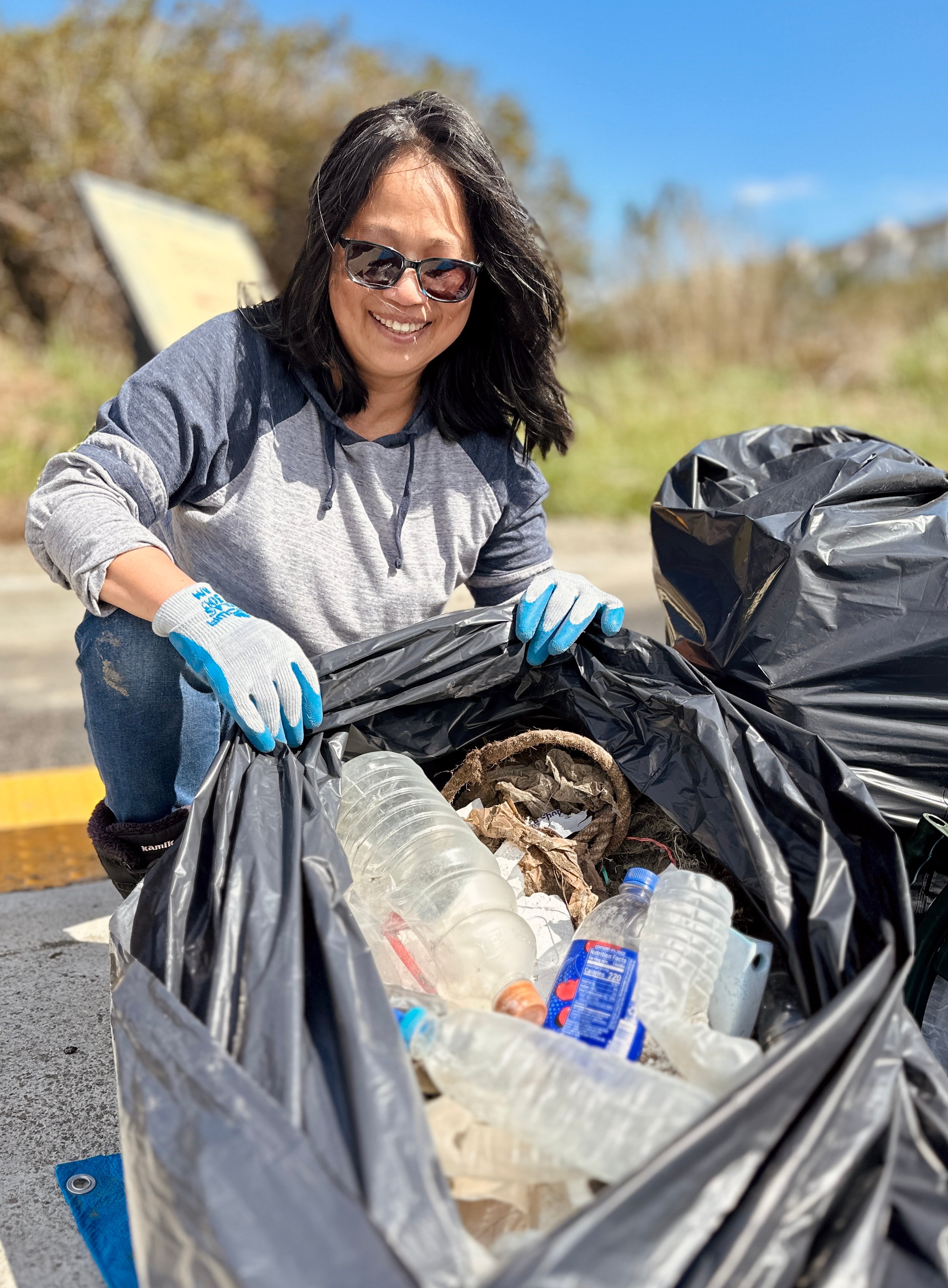 A volunteer poses in front of a trash bag full of single use plastic bottles and other beach trash 