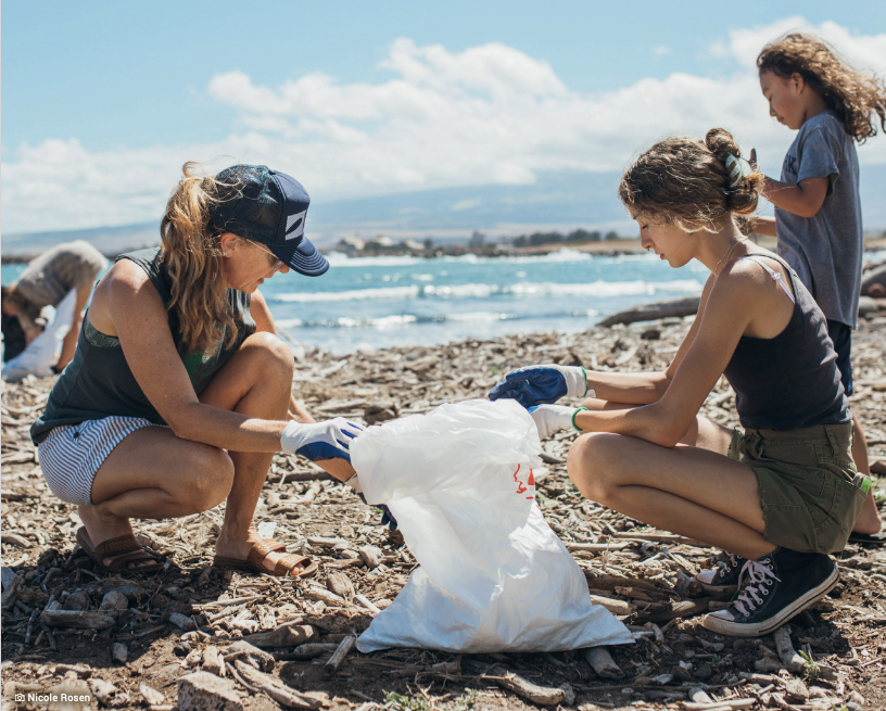 Three individuals participate in a beach cleanup, with two crouching holding a garbage bag, sifting through driftwood debris to remove man-made litter