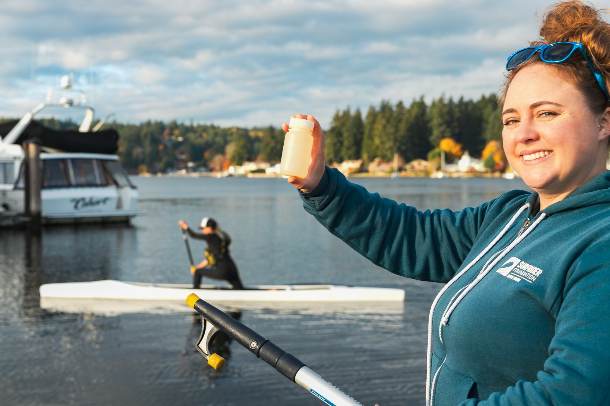 A female volunteer collecting water samples while a kayaker enjoys recreating on the water