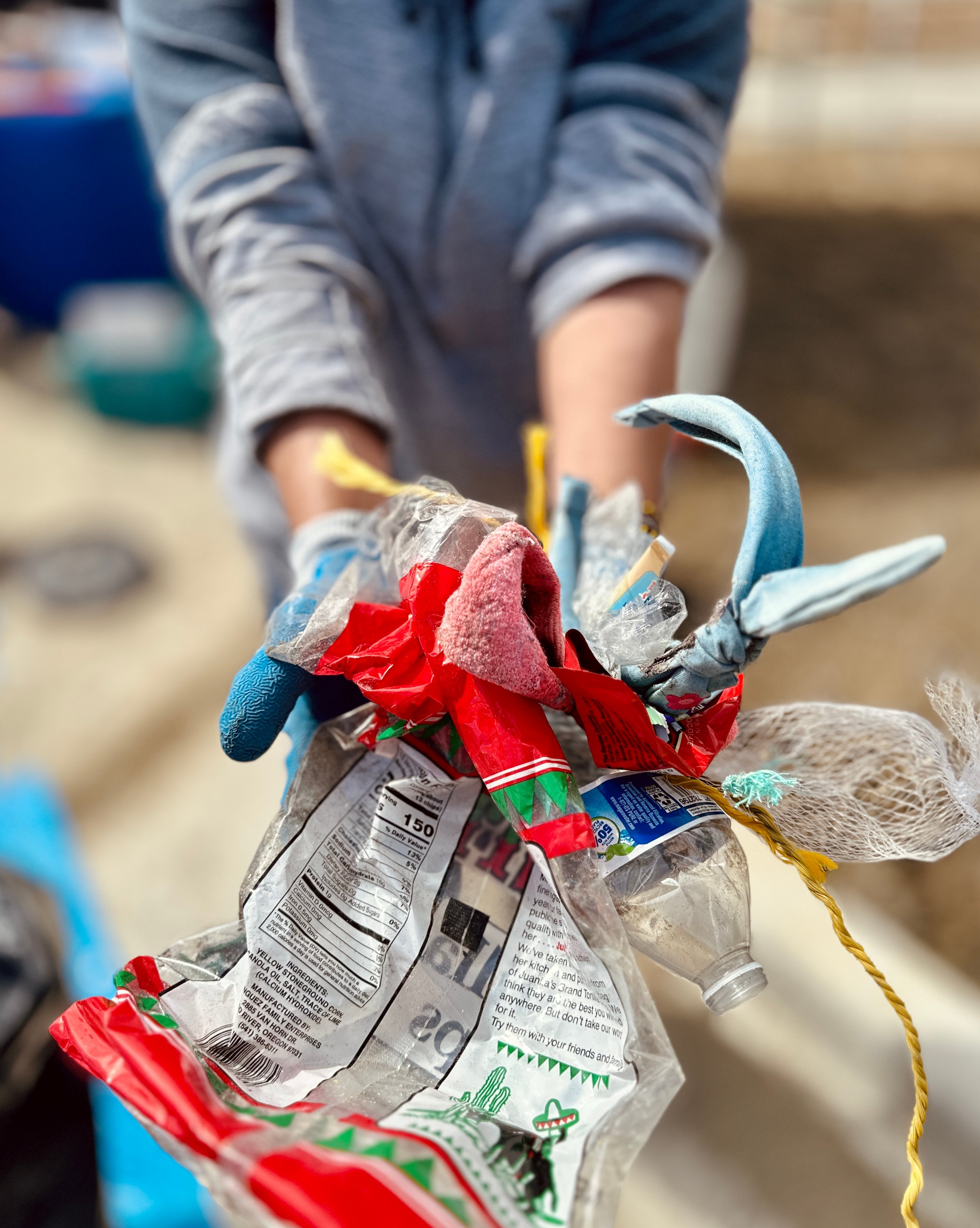 A volunteer holds up a handful of trash removed from the beach, including a chips bag, single-use water bottle, aquaculture rope, and a kid's headband